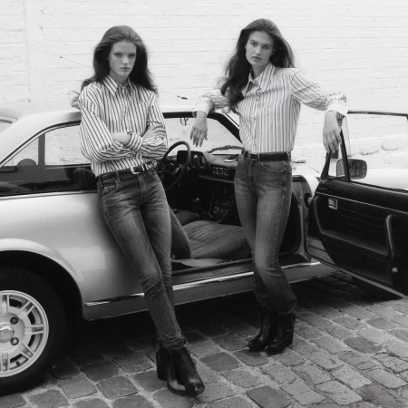 Two women pose by a vintage car on a cobblestone street. They wear jeans, striped shirts, and boots in a black-and-white editorial shot.