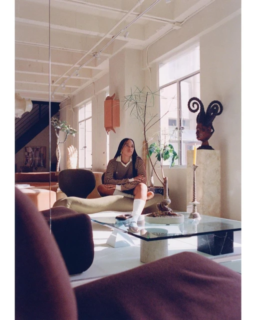 Woman in striped shirt sits on green couch in bright, modern living room with sculptures, plants, glass table, and large windows.