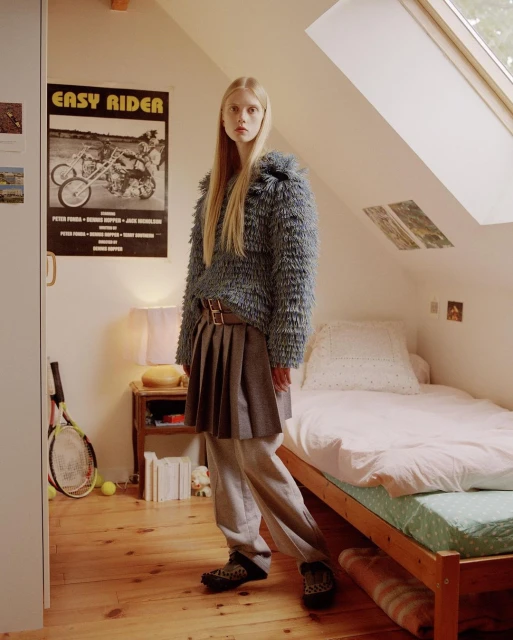 A slender blonde fashion model poses in an attic room wearing a blue textured sweater, pleated skirt, sneakers, and Easy Rider poster in background.