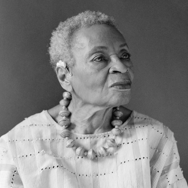 Black and white portrait of a mature African American woman with short grey hair, wearing a beaded necklace and white top, looking upwards.