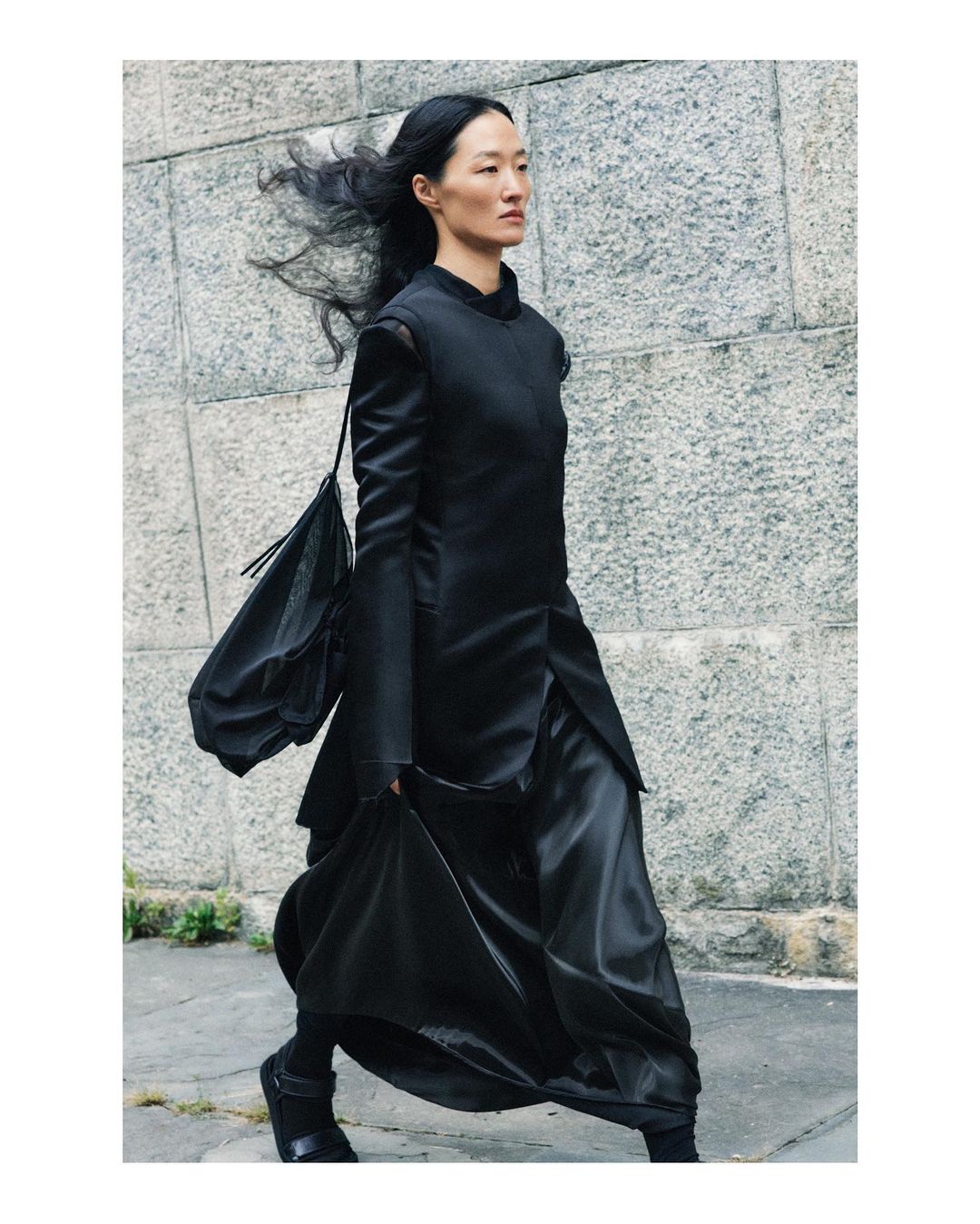 Asian woman in stylish black outfit walking outdoors against a textured wall in a fashion context, showcasing modern elegance.