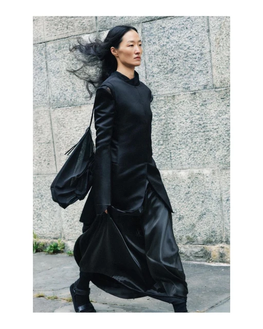 Asian woman in stylish black outfit walking outdoors against a textured wall in a fashion context, showcasing modern elegance.