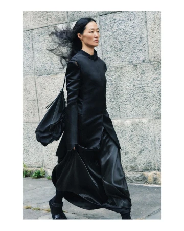 Asian woman in stylish black outfit walking outdoors against a textured wall in a fashion context, showcasing modern elegance.