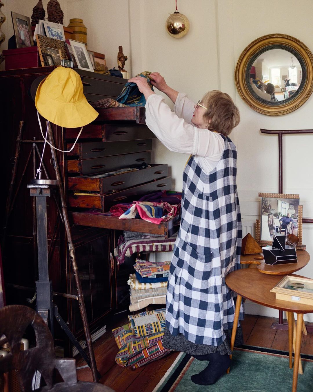 Woman arranging textiles in vintage cabinet with open drawers in a well-lit interior with antique decor and furnishings.