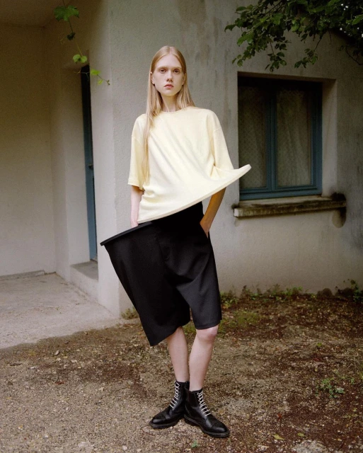 A slender, fair-skinned young woman models a light yellow t-shirt, dark, baggy shorts and lace-up boots in front of a stucco wall.