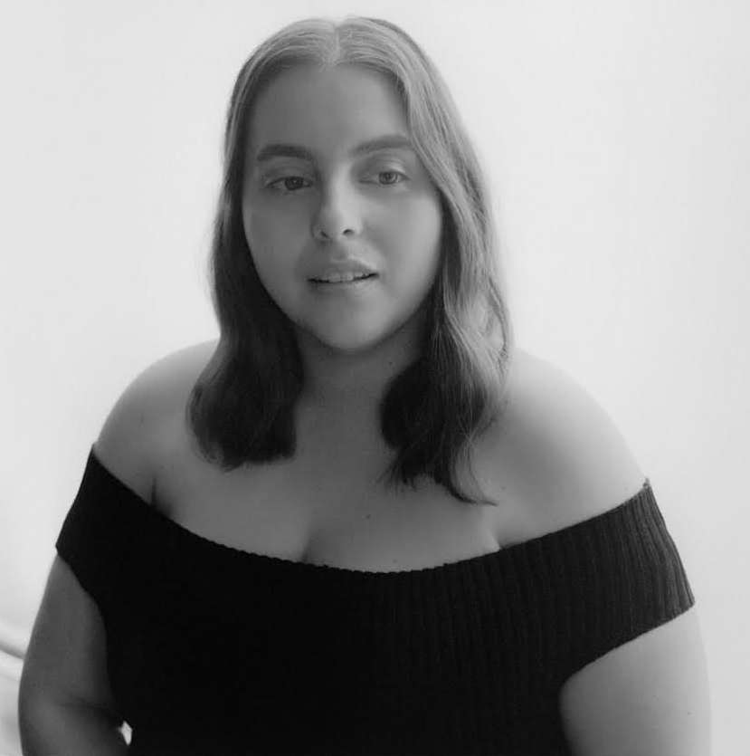 Portrait of a young woman with wavy shoulder-length hair wearing a black off-the-shoulder top against a bright, neutral background.
