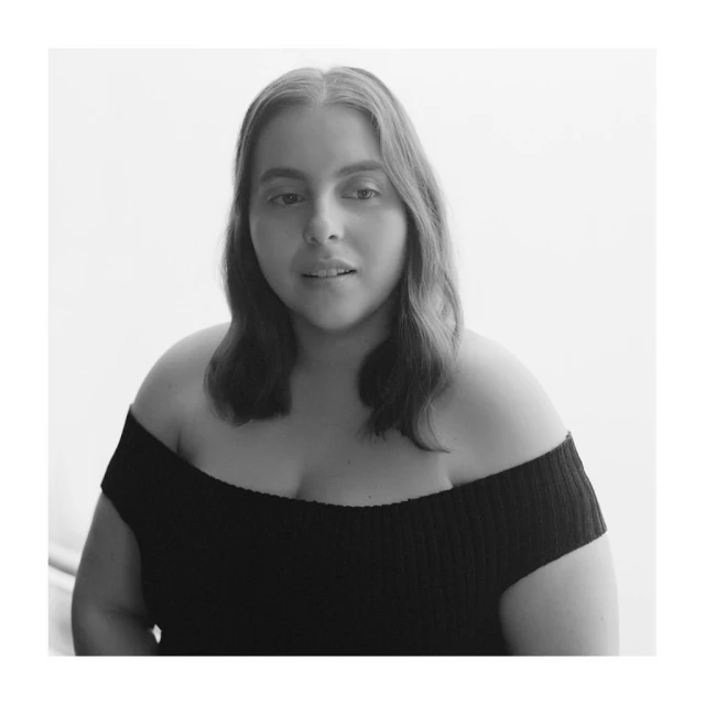 Monochrome portrait of a fair-skinned woman with shoulder-length hair wearing a black off-the-shoulder top against a bright, plain background.