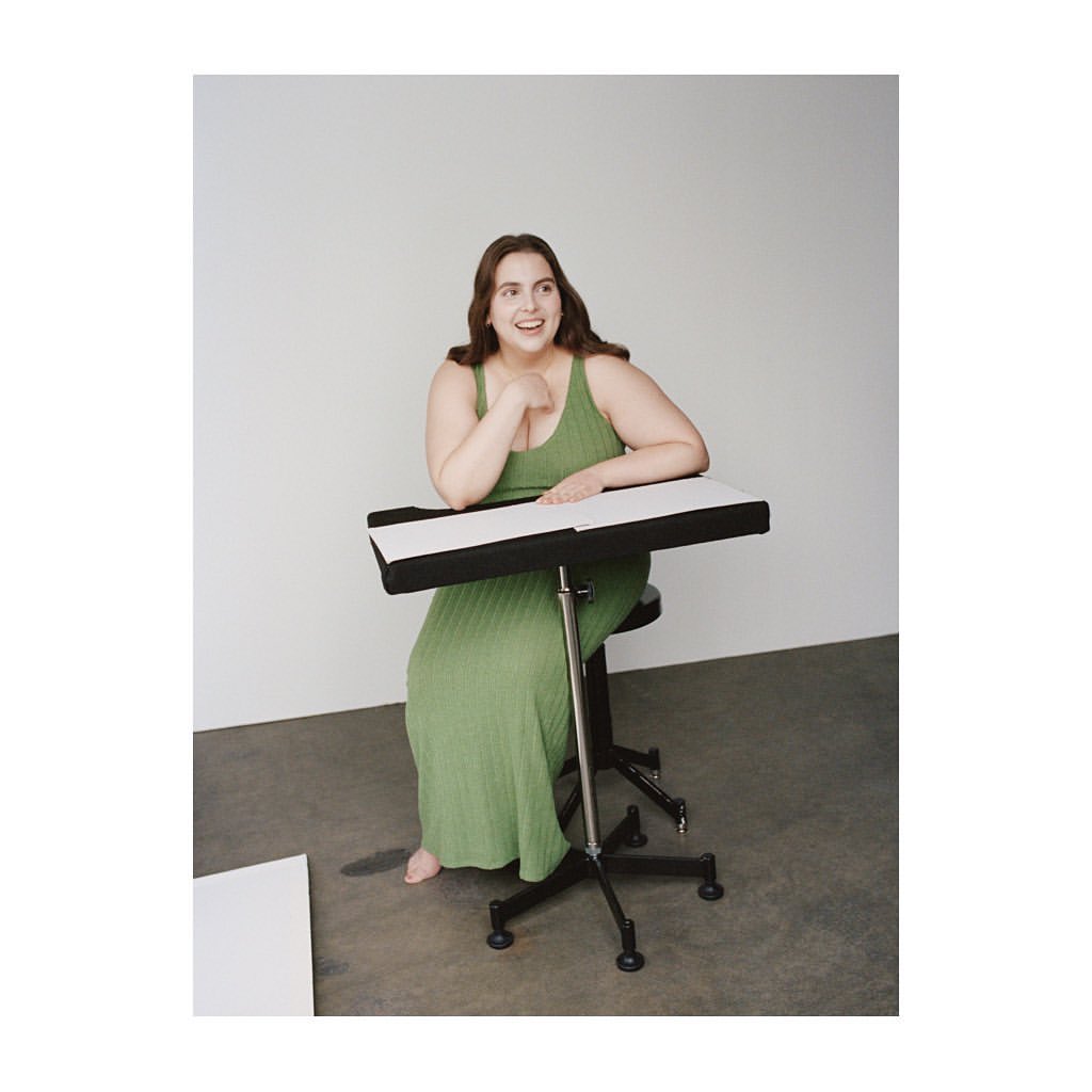 A smiling woman in a green dress sits at an adjustable drafting table in a studio with a blank sheet of paper on the floor.