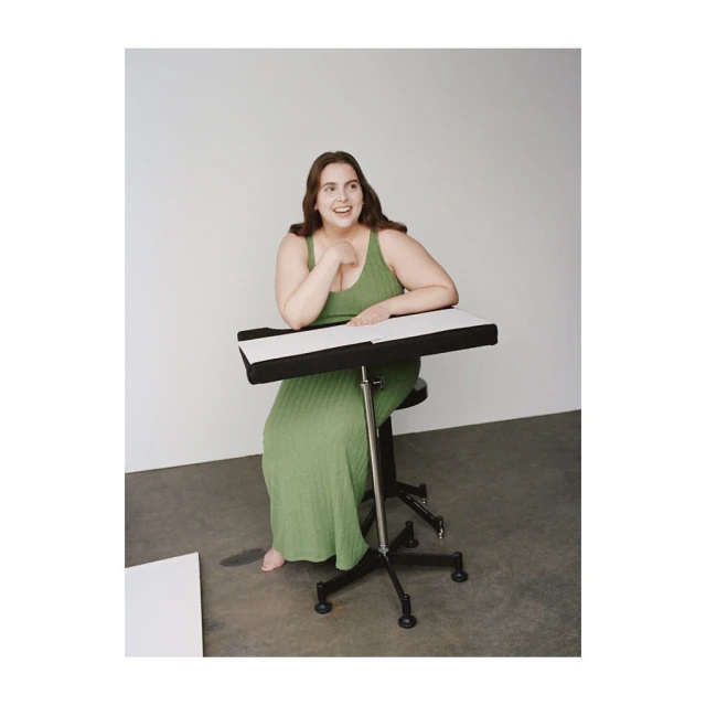 A smiling woman in a green dress sits at an adjustable drafting table in a studio with a blank sheet of paper on the floor.