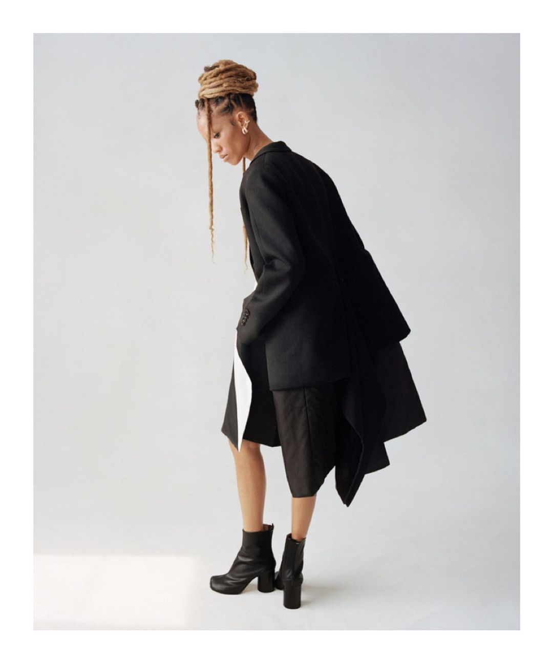 Fashionable woman in oversized black blazer, neutral mini-skirt, and black boots poses in a studio with bright lighting and a white backdrop.