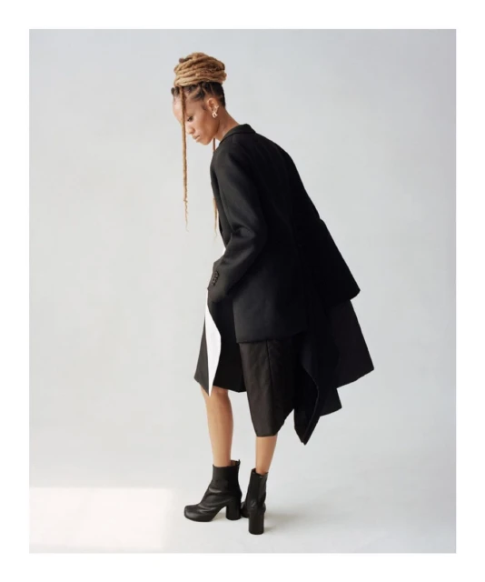 Fashionable woman in oversized black blazer, neutral mini-skirt, and black boots poses in a studio with bright lighting and a white backdrop.
