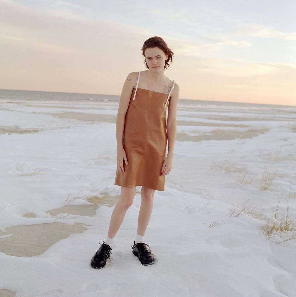 Young woman in brown dress poses on snowy beach. Minimalist fashion shoot in natural light with a cold, wintry feel, distant ocean.