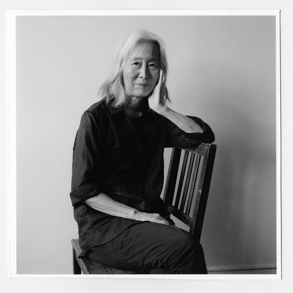 Portrait of an elderly woman with shoulder-length white hair seated in a wooden chair against a neutral background in black and white.