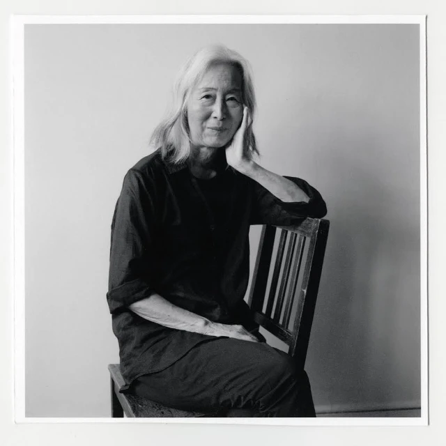 Portrait of an elderly woman with shoulder-length white hair seated in a wooden chair against a neutral background in black and white.