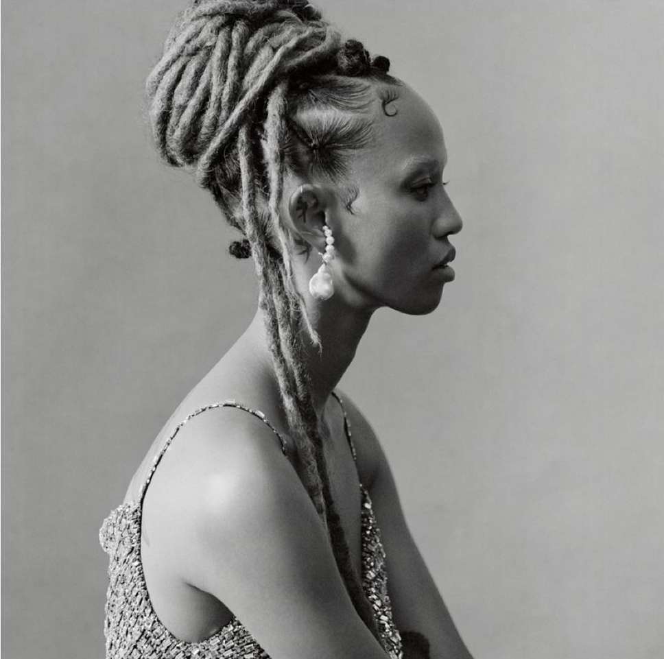 Portrait of a black woman with styled dreadlocks, wearing a sparkly top and large baroque pearl earrings in a studio setting.