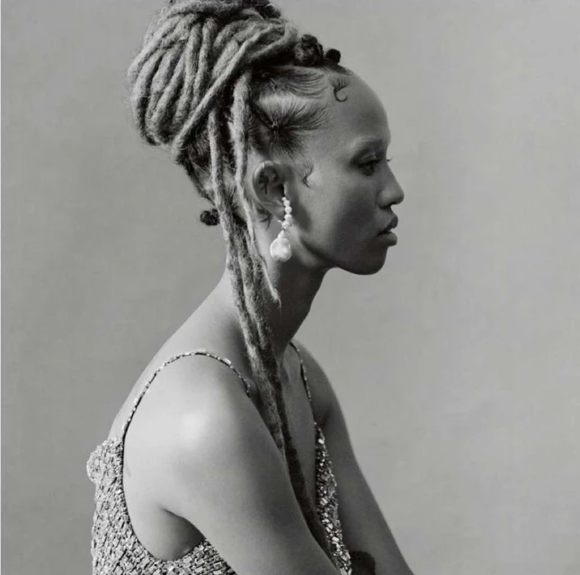 Portrait of a black woman with styled dreadlocks, wearing a sparkly top and large baroque pearl earrings in a studio setting.