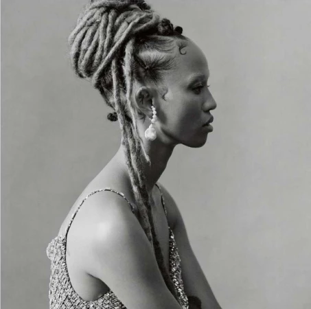 Portrait of a black woman with styled dreadlocks, wearing a sparkly top and large baroque pearl earrings in a studio setting.