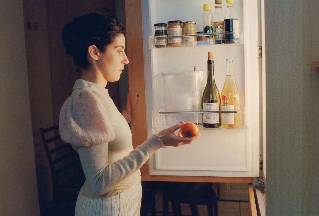 Young adult woman in white outfit holds peach in front of open refrigerator with wine bottles, food jars, indoor setting.