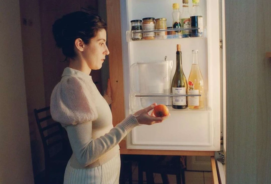 Young adult woman in white outfit holds peach in front of open refrigerator with wine bottles, food jars, indoor setting.