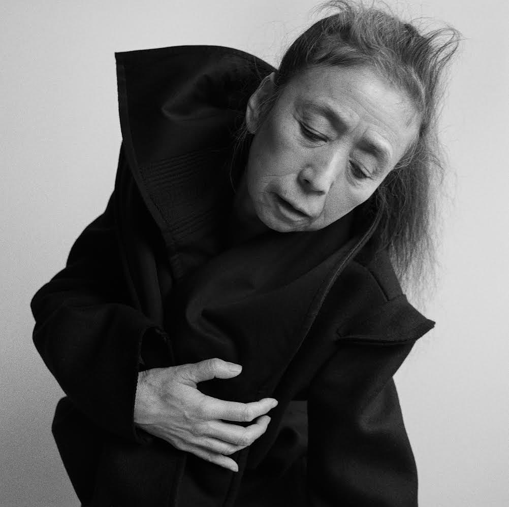 Monochrome portrait of an adult female in a dark coat with a large collar, angled light, and a plain background.