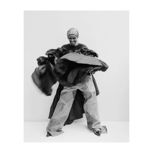Monochromatic portrait of a Black woman in motion, wearing a headscarf, oversized jeans, and a dark, flowing garment in a studio setting.