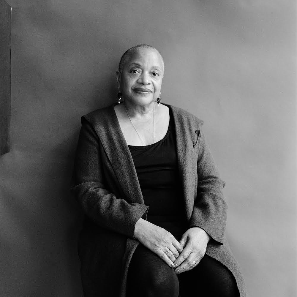 Portrait of an older black woman seated against a plain grey background, wearing a dark coat and pendant necklace.