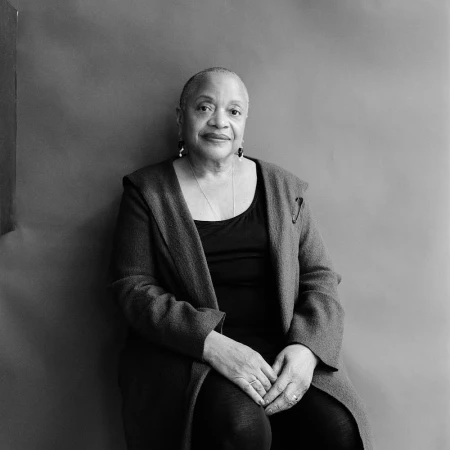 Portrait of an older black woman seated against a plain grey background, wearing a dark coat and pendant necklace.