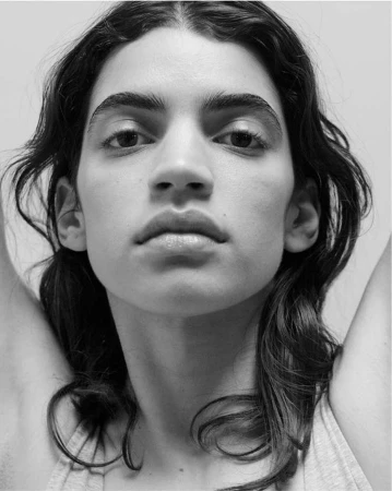 Close up portrait of a person with dark wavy hair and a light tank top against a plain light background photographed by Jody Rogac