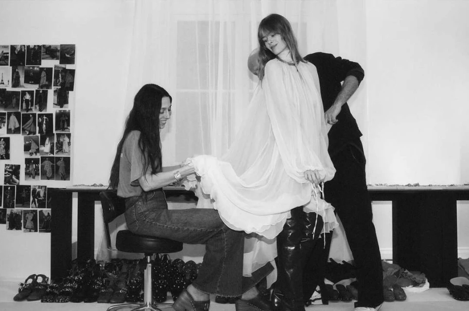 Black and white photograph of a fashion fitting with a model wearing a white dress, designer, and a stylist in a photo studio.