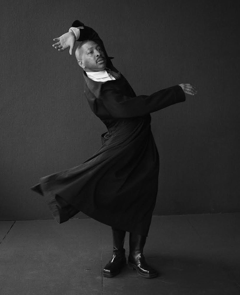 Monochrome portrait of a man in a dark, flowing coat dancing against a gray wall, capturing motion and style in a studio setting.