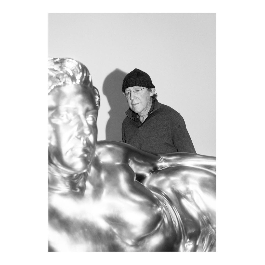 Black and white portrait of an adult male wearing a beanie and sweater next to a metallic statue in an indoor setting.