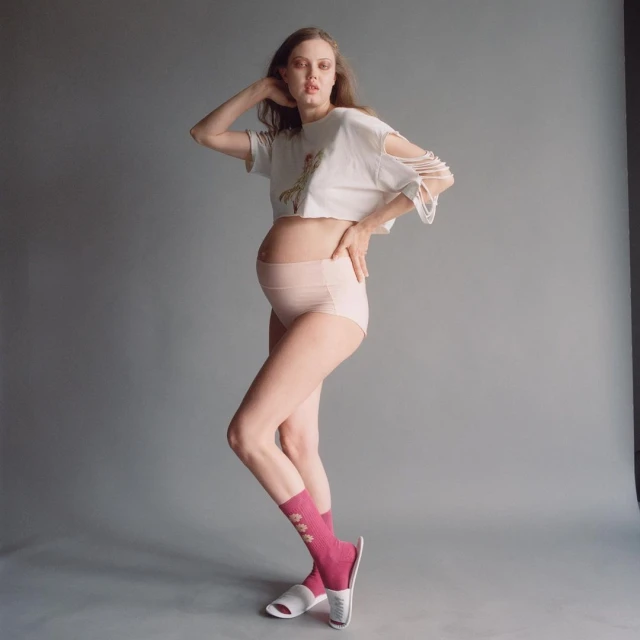 A pregnant woman poses in a photo studio wearing a t-shirt, briefs, socks, and slippers against a grey backdrop.