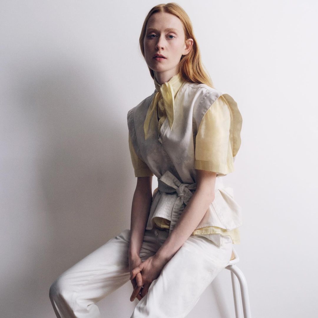 Elegant red-haired adult female sits on white stool wearing white pants, linen vest, and soft yellow scarf in a minimalist photo studio.