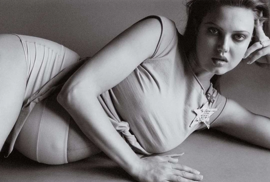 Monochrome portrait of a young woman reclining on the floor, wearing a light-colored top and bottoms in a photo studio setting.