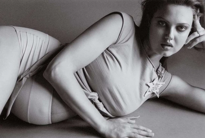 Monochrome portrait of a young woman reclining on the floor, wearing a light-colored top and bottoms in a photo studio setting.