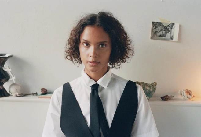 Portrait of a woman with curly hair wearing a white collared shirt, black tie, and black vest against a white wall.