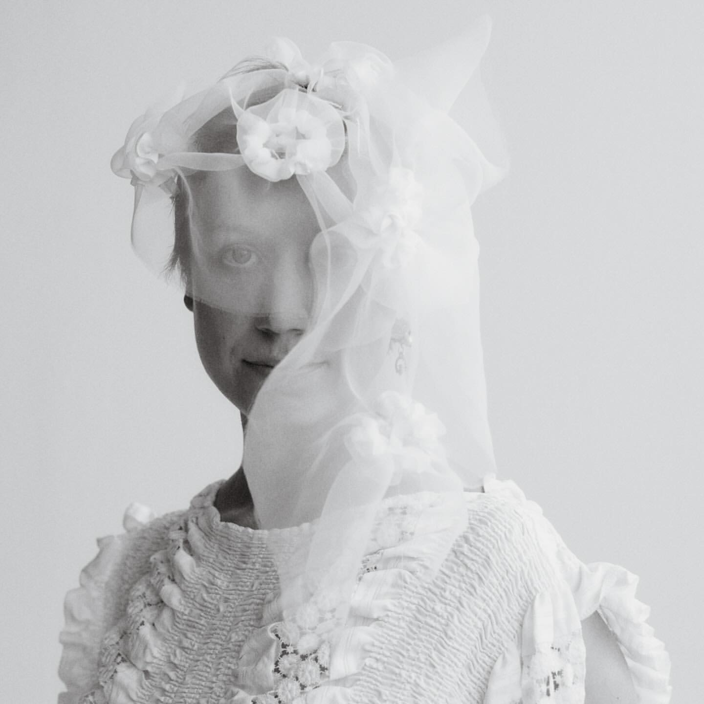 Monochrome portrait of a person in a white dress and veil with floral embellishments against a pale grey background.