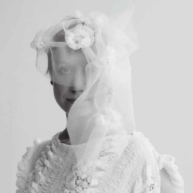Monochrome portrait of a person in a white dress and veil with floral embellishments against a pale grey background.