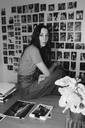 Monochrome portrait of a young woman sitting on a wooden table, surrounded by photos and flowers, against a wall covered in pictures.