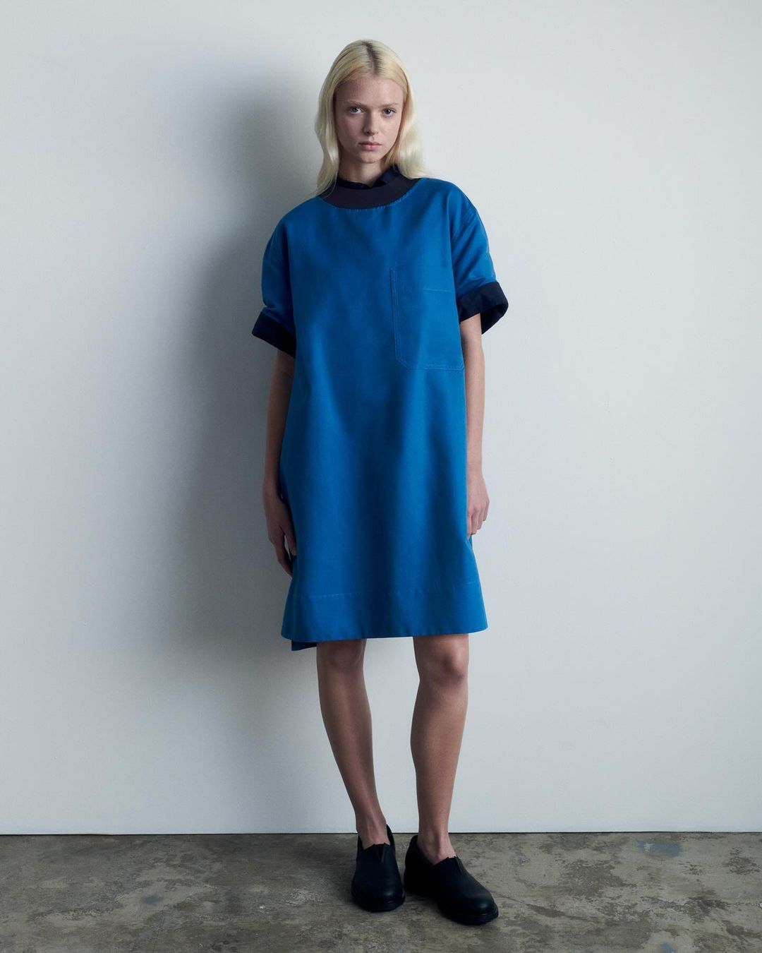 A fair-skinned adult woman stands in a blue dress and black shoes against a white wall, facing the camera in a studio setting.