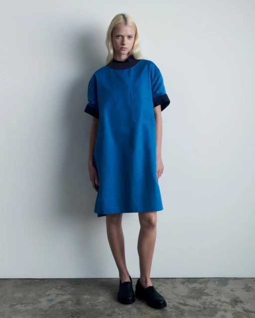 A fair-skinned adult woman stands in a blue dress and black shoes against a white wall, facing the camera in a studio setting.