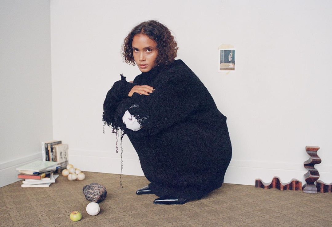 Woman with curly hair in a black oversized sweater crouching in an indoor setting with books, stone, balls, and wooden forms.