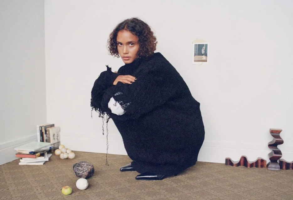 Woman with curly hair in a black oversized sweater crouching in an indoor setting with books, stone, balls, and wooden forms.