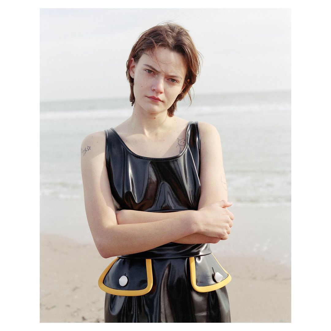 Young woman with short hair poses in a shiny black and yellow dress on a beach, with a soft, natural lighting and ocean background.