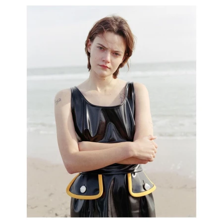 Young woman with short hair poses in a shiny black and yellow dress on a beach, with a soft, natural lighting and ocean background.