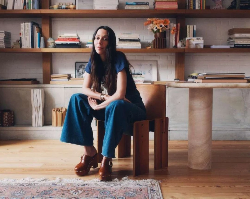 A woman with long dark hair wearing a blue top and denim pants is sitting on a wood chair indoors with a bookshelf in the background.