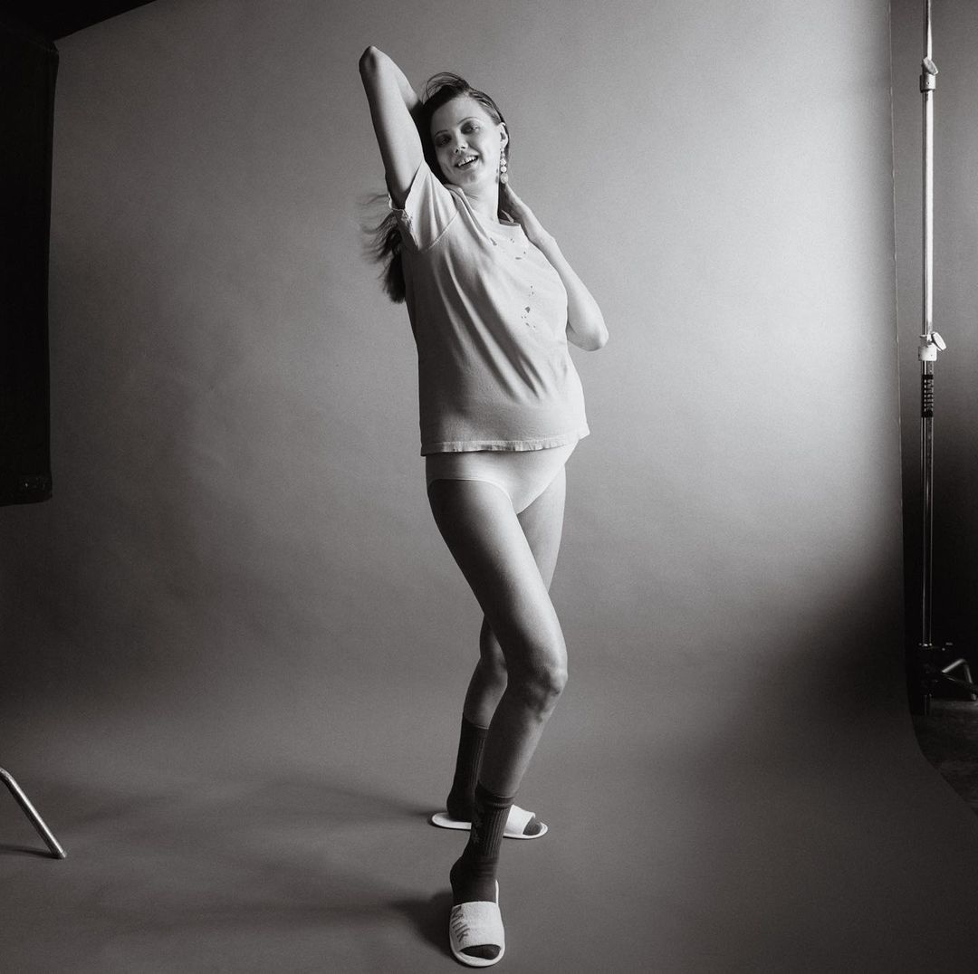 Pregnant woman in t-shirt and underwear posing in a studio with grey seamless backdrop, photographed by Jody Rogac.
