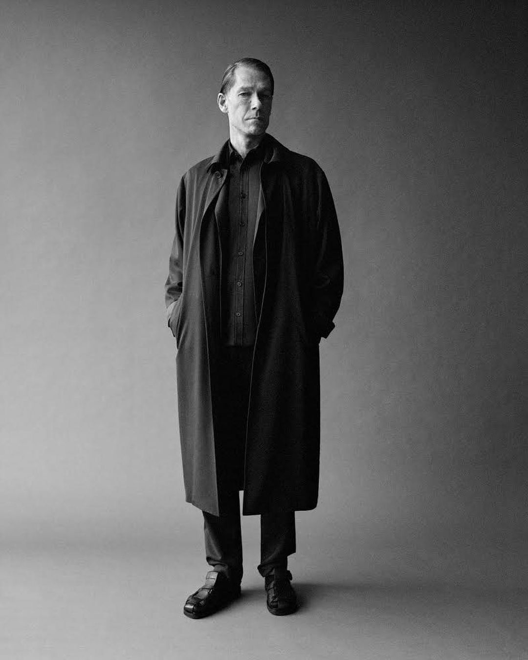 Monochrome portrait of a man in a long dark coat standing against a plain grey studio backdrop.