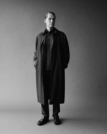 Monochrome portrait of a man in a long dark coat standing against a plain grey studio backdrop.