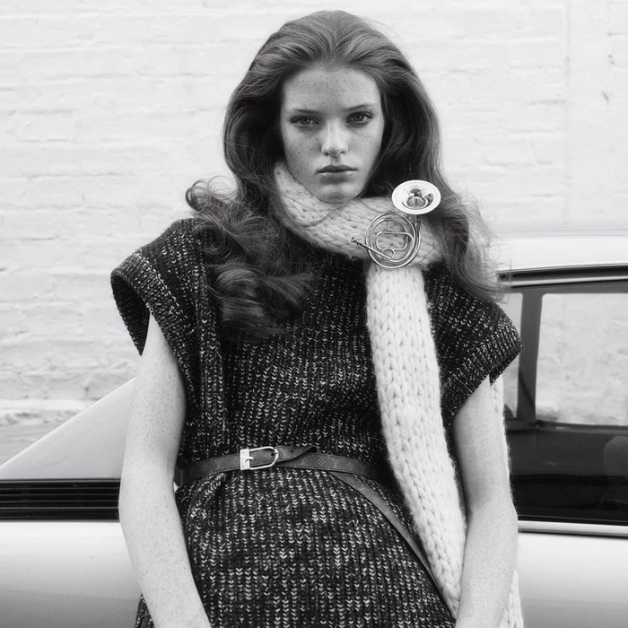 Model with long wavy hair wearing a knitted scarf and patterned dress leaning against a vintage car